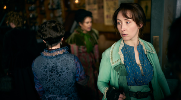 Erin Doherty (early 30s, light skin tone) is in character as Mary Carr, dressed in victorian clothing with a turquoise coat and skirts. She looks off camera at something we cannot see. Behind her are two other female actors, both dressed similarly to her 