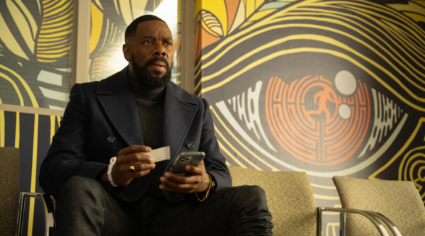 Colman Domingo, a black man in is fifties, sits indoors in a navy blue peacoat-style waist-length jacket, holding a phone and small piece of paper