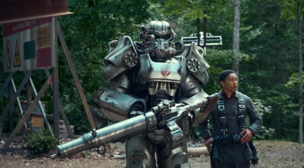 Two people walk in a forest, one in a large chrome mec suit