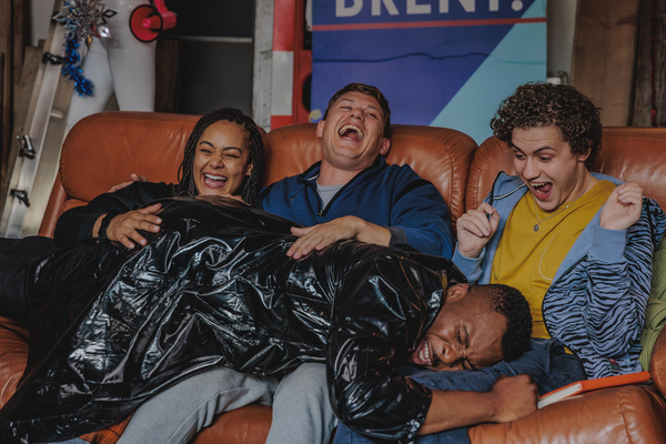 A group of four friends sit on a rust coloured sofa, laughing as one of them lies on their laps