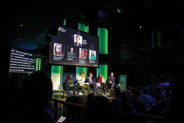 A panel of 3 men and two women sit on a stage in front of a screen with their pictures and names