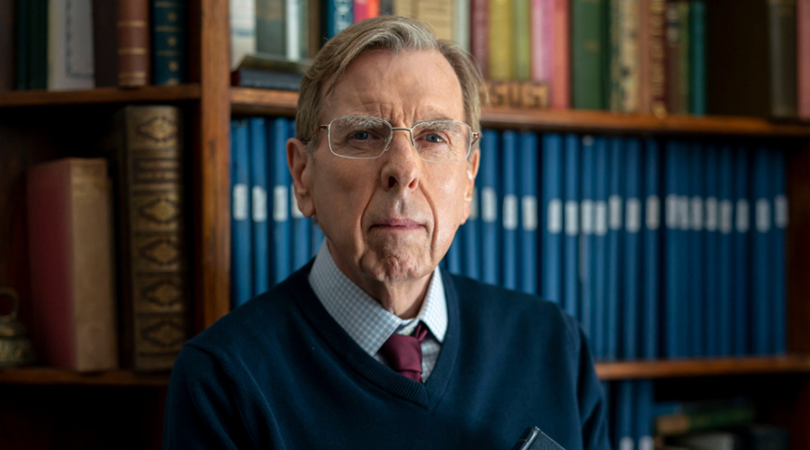 Timothy Spall stands in front of bookshelves, wearing glasses and looking pensive