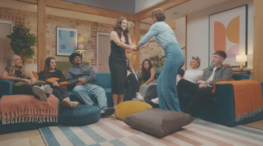 Two woman join hands, facing each other and smiling, while seven other people sit on sofas, looking up at them happily