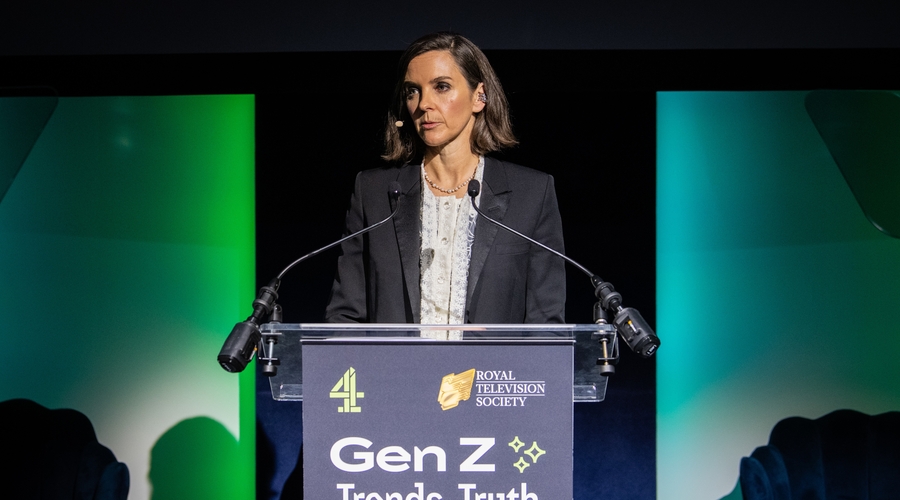 A woman in her 50s with dark hair and wearing a black blazer and white shirt stands at a lecturn covered with the words: 'Gen Z: Trends, Truth and Trust'