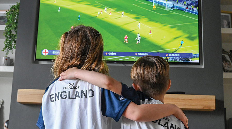 Two kids wearing England football shirts watch England play in Euro 2024 on TV 