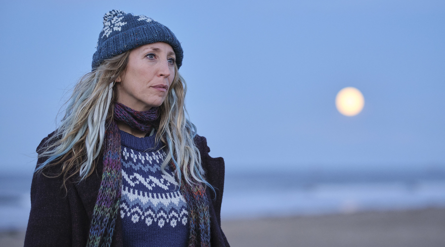 a woman in her 40s faces to the right, standing on a beach with wind in her hair, which is blonde with blue highlights. She wears a blue jumper, hat and a multicoloured scarf. She appears troubled but hardened.