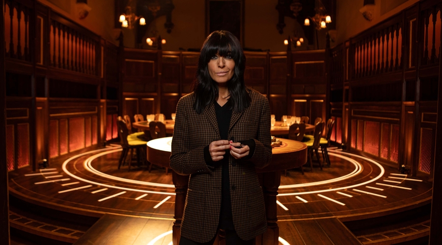 Claudia Winkleman, a woman with a light skin tone, and dark hair with a long fringe, stands in front of a large round table surrounded by seats