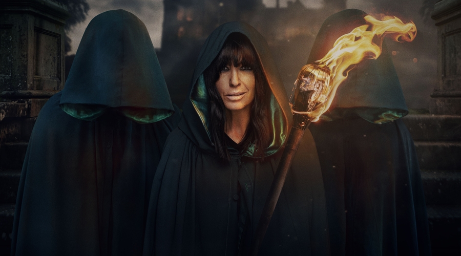 Claudia Winkleman stands in the middle with a green cloak holding a fire on a stick, with two figures either side of her, covered by their hoods