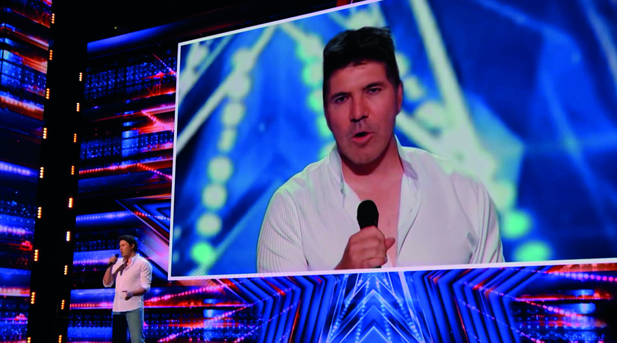 A white man sings on the America's Got Talent stage, but is shown on the big-screen behind him as Simon Cowell