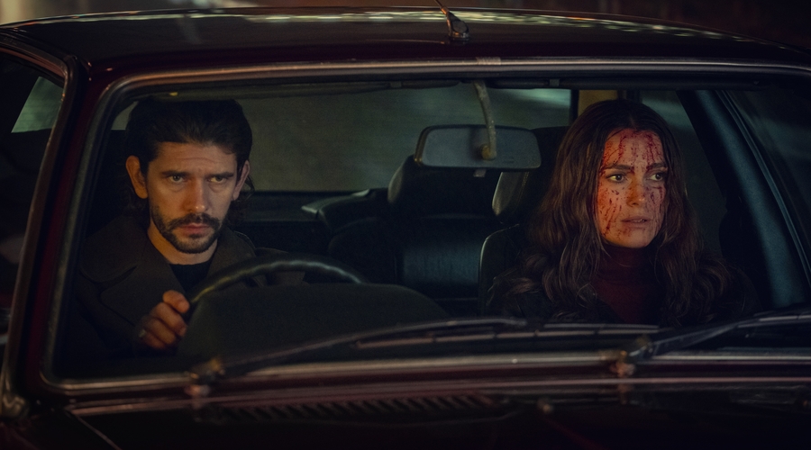 Ben Whishaw and Keira Knightley, both in their late 30s/early 40s, sit side by side in a car. Both have light skin tones, Ben has dark hair and a beard. Keira has long light brown hair that's waved, and she is covered in blood.