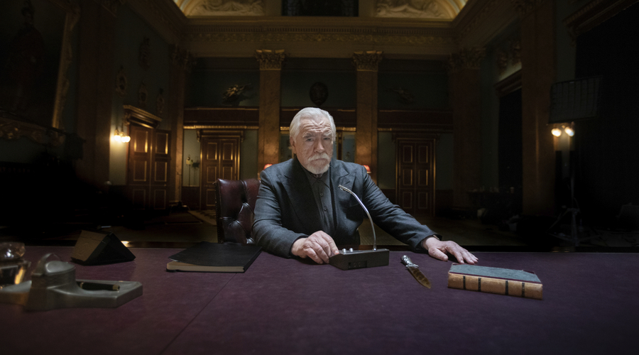 Brian Cox sits at a desk in an opulent office, ready to speak into a microphone