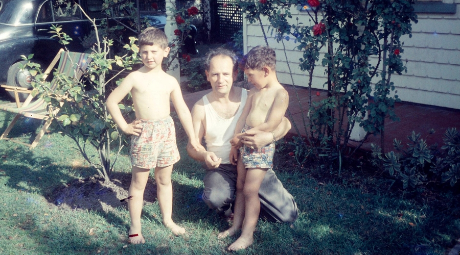 Two boys stand either side of their dad in an old, slightly blue-tinted family photo, all of them stood on a lawn