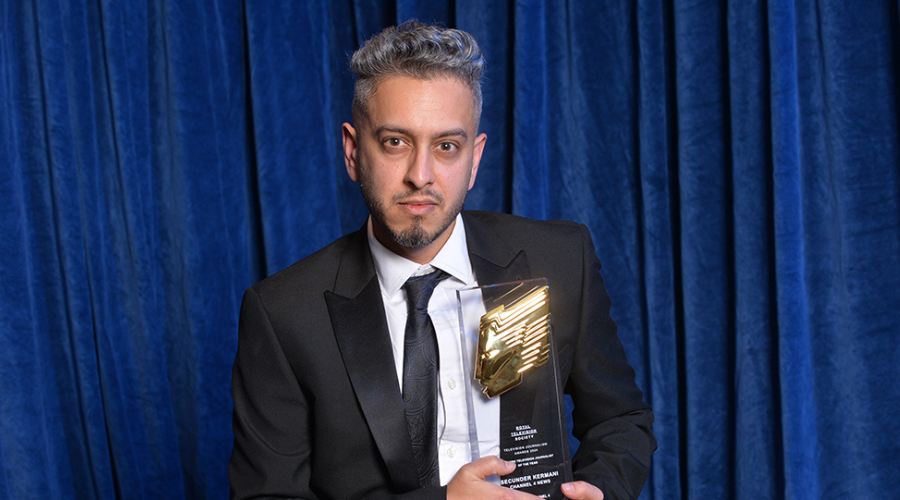 Secunder Kermani sits in a suit holding an RTS Award, looking into the camera in front of a blue curtain backdrop