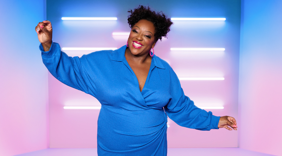 Judi Love smiles to the camera in a blue dress, in front of a pastel blue and pink backdrop, which features white horizontal neon lights