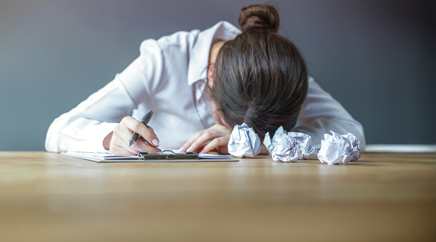 A woman sits with her head on a desk, next to scrunched up bits of paper