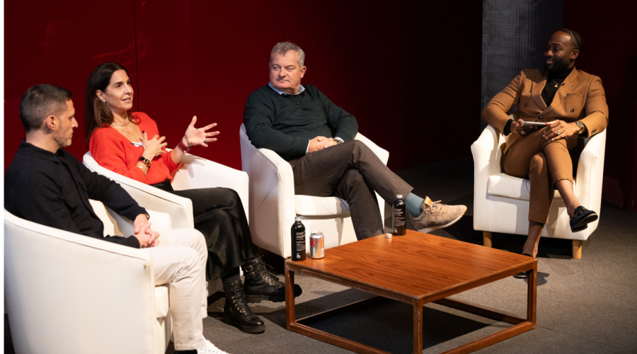 Four individuals seated on stage on a panel: a man with short hair in a black top and light trousers, a woman with dark long hair in a red blouse, black skirt and boots, a man with grey hair in a dark jumper and brown trousers, and a man in a brown suit