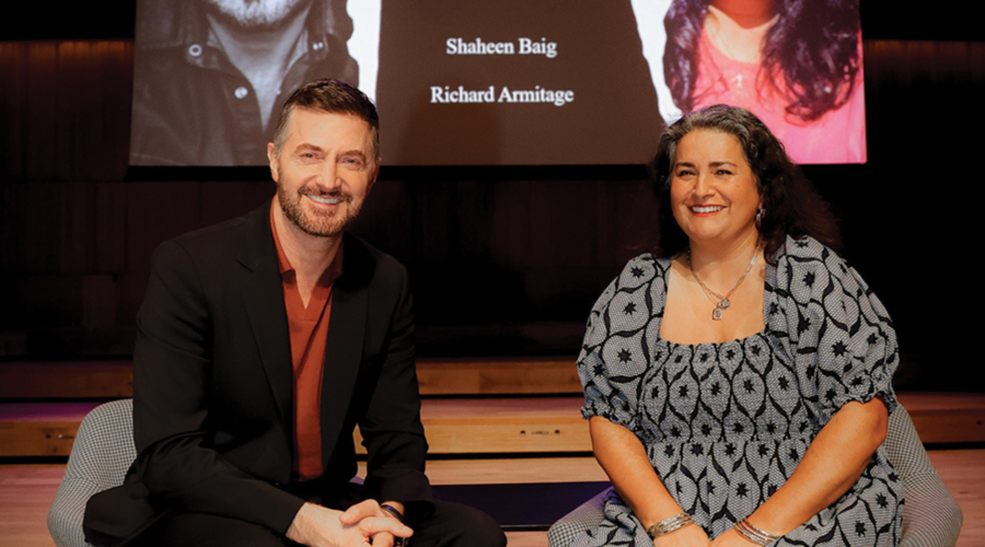 Shaheen Baig and Richard Armitage smile into the camera, sat down in front of a projector screen showing both their names