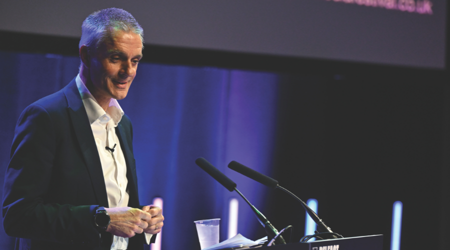 Tim Davie, who has short white hair and is in his mid-fifties, stands at a podium, smiling slightly