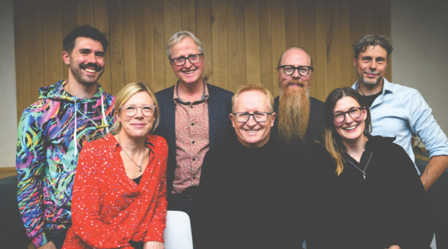 The seven people stand indoors in front of wood panelling, smiling to the camera