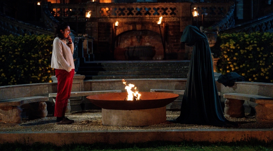 A white woman with dark hair stands by a firepit outside a castle and opposite a fully cloaked person