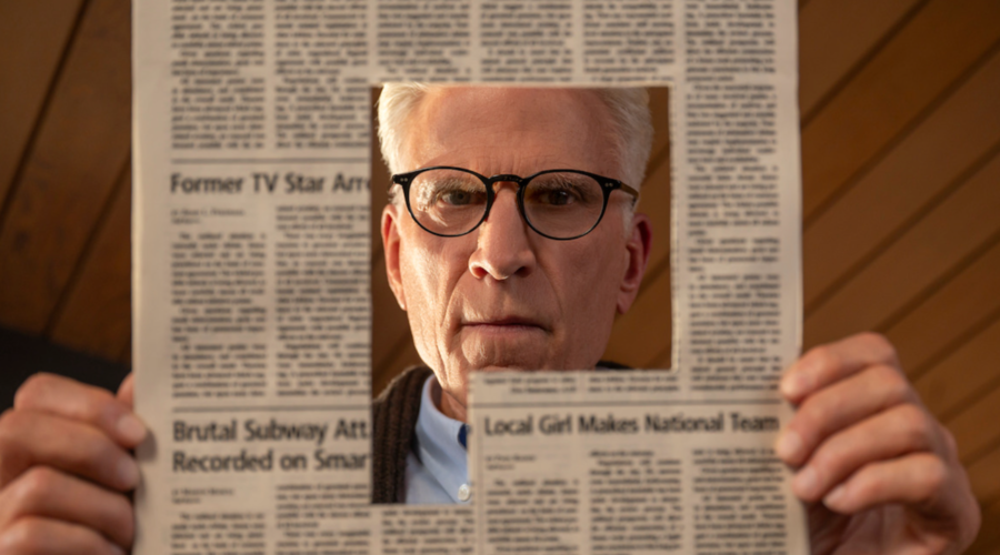 Ted Danson, a man in his 70s wearing black glasses, looks down at the page of a newspaper with a large article cut out of it, allowing us to see his face peering through