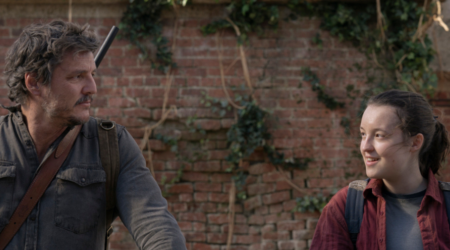 Pedro Pascal, a Chilean-American man in his forties, and Bella Ramsey, a person in their late-teens in the image, stand side by side, looking at each other in front of a brick wall