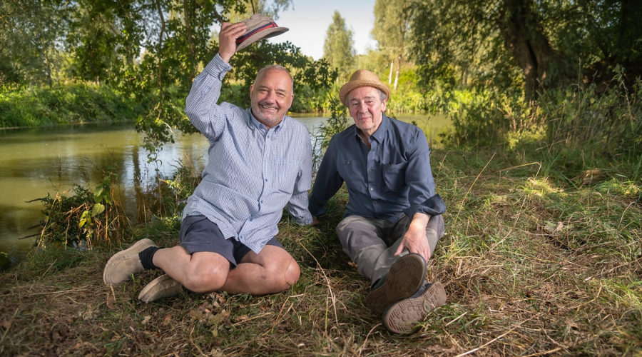 Bob Mortimer and Paul Whitehouse sit next to each other by a lake, looking into the camera and smiling, with Mortimer taking his hat off in friendly greeting