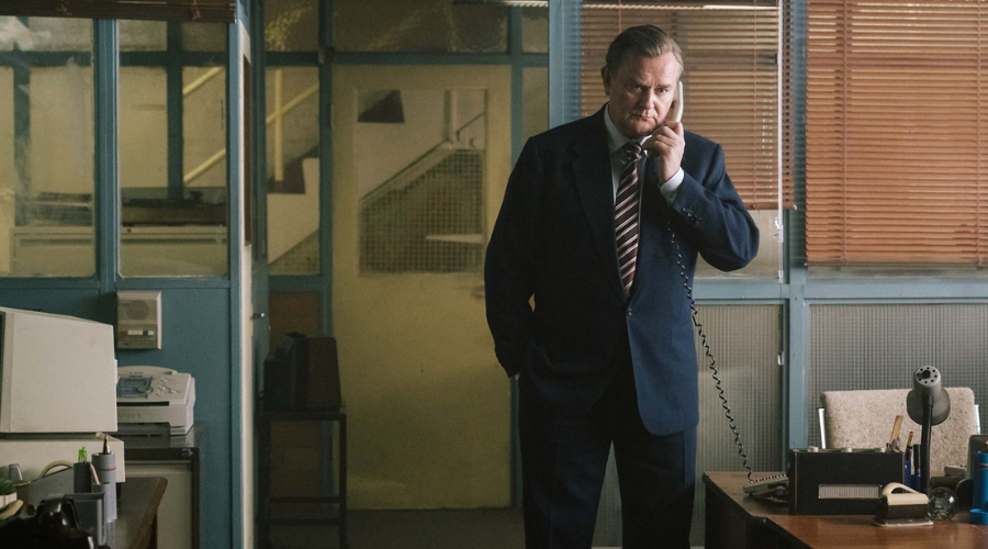 Hugh Bonneville, a white man in his sixties, stands in an office on a phone with a cord, looking concerned