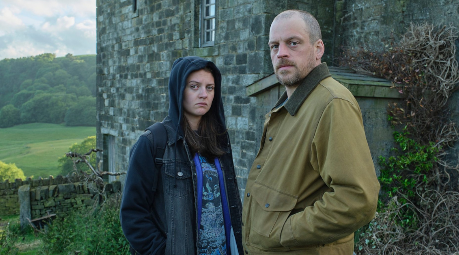 The two stand outdoors in front of a grey stone building, behind which is an expansive green field. Neither of them smile.