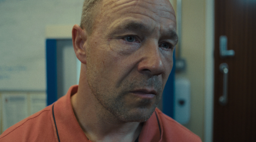 Stephen Graham, a white man in his fifties, looks into the middle distance in an extreme close-up, looking stressed, wearing an orange polo shirt