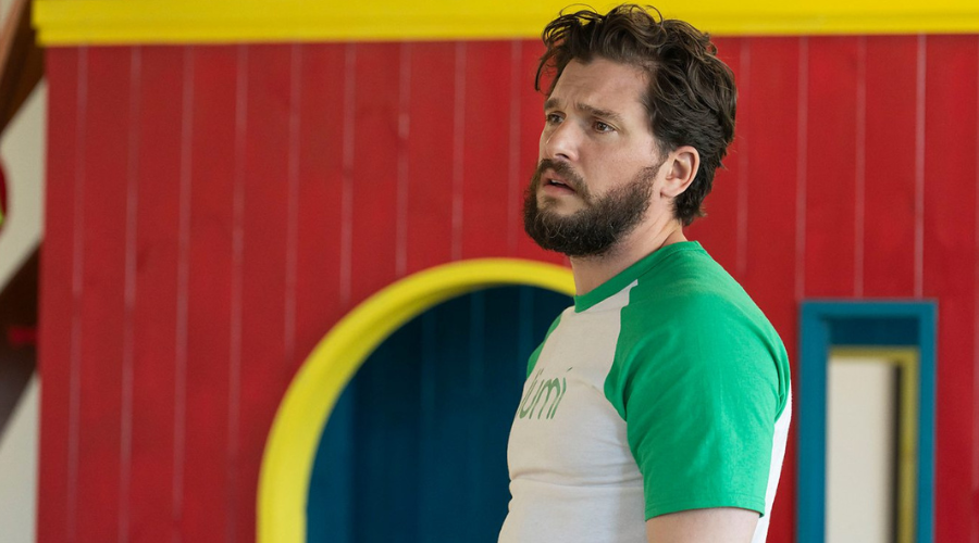 Kit Harington stands indoors, looking crestfallen in a white and green t-shirt with the logo for Lumi, an in-world green energy company 