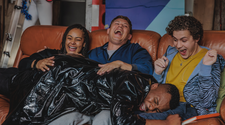 A group of four friends sit on a rust coloured sofa, laughing as one of them lies on their laps