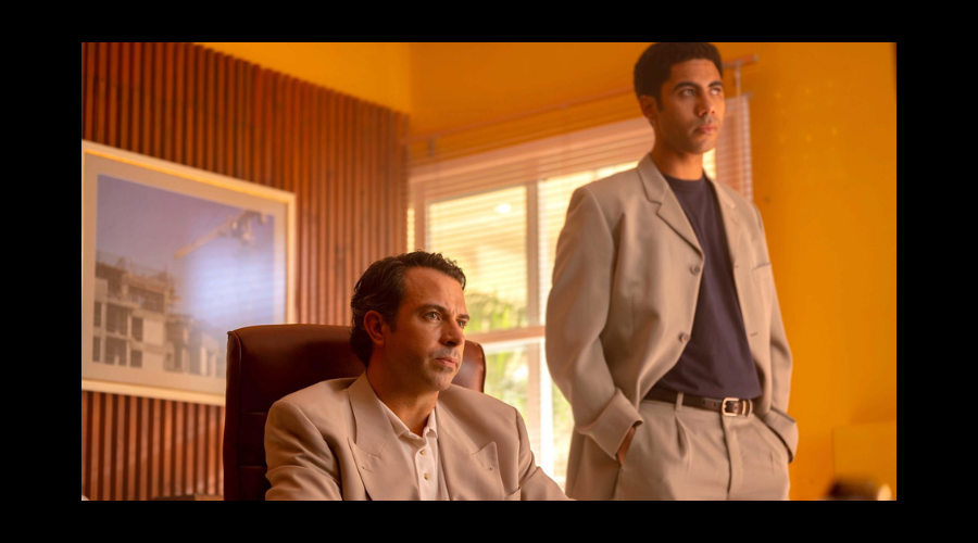 Tom Cullen, a white man in his thirties, sits next to Joshua Samuels, a young Black man, who stands, the two of them looking at something off-camera