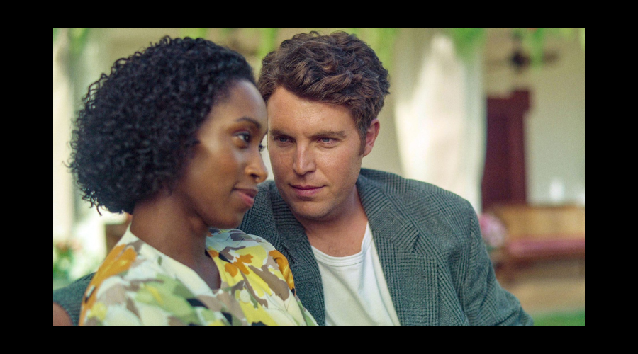 Rochelle Neil, a Black woman in her thirties, sits next to Tom Hughes, a white man in his thirties. The two smile slightly, Tom looking at Rochelle, and Rochelle looking into the middle distance