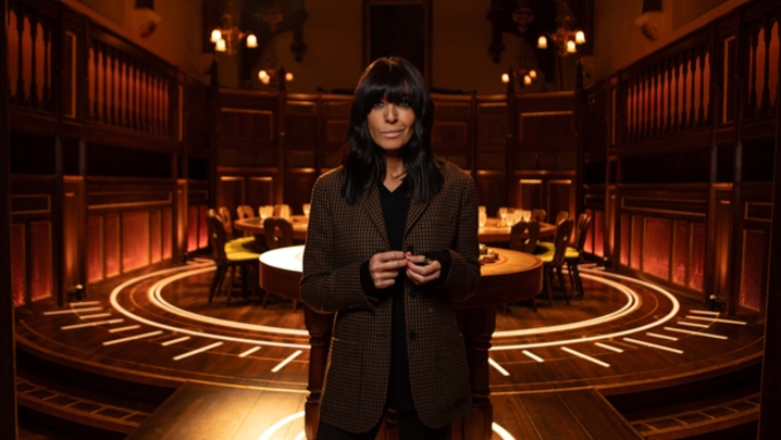 Claudia Winkleman, a woman with a light skin tone, and dark hair with a long fringe, stands in front of a large round table surrounded by seats