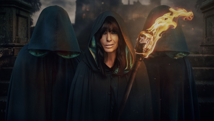 Claudia Winkleman stands in the middle with a green cloak holding a fire on a stick, with two figures either side of her, covered by their hoods