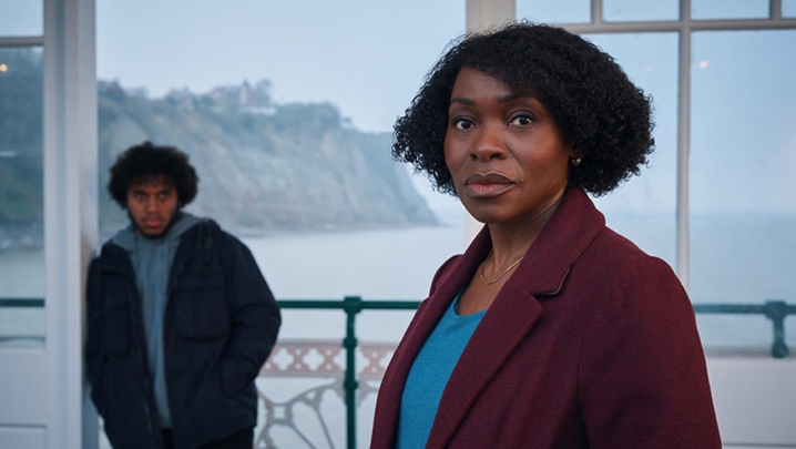 A black woman in a red coat stands near the sea with a black man in a black coat in the background