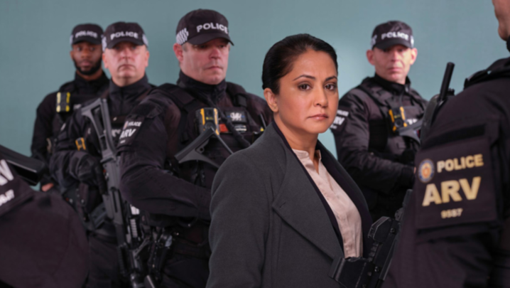A woman in a grey coat stands in front of a group of armed police