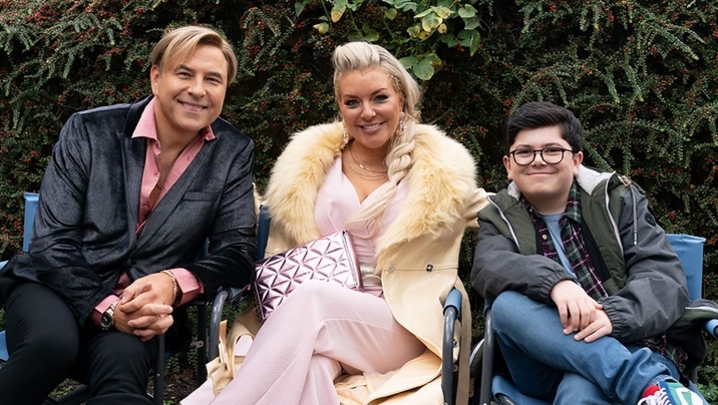 David Walliams sits in a black suit and pink shirt, next to him Sheridan Smith sits in a pink jumpsuit and silver jewellery, next to her Archie Yates wears glasses and sits in jeans a jacket and trainers