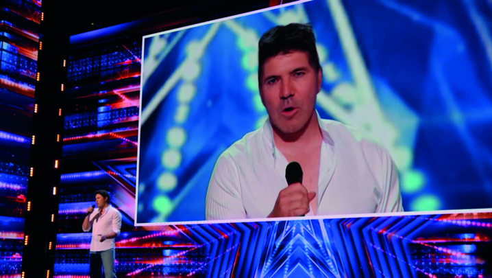 A white man sings on the America's Got Talent stage, but is shown on the big-screen behind him as Simon Cowell