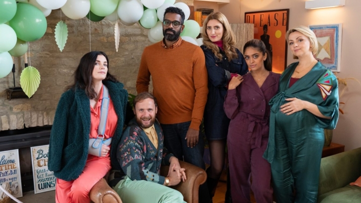 Aisling Bea, Colin Hoult, Romesh Ranganathan, Jessica Knappett, Mandeep Dhillon and Lisa McGrillis sit and stand around a chair in a living room, which is decked out with balloons