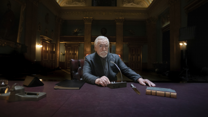 Brian Cox sits at a desk in an opulent office, ready to speak into a microphone