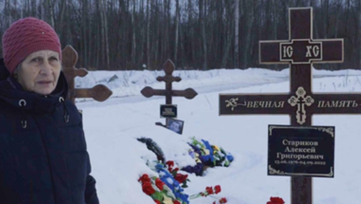 An elderly woman stands in front of three graves, which have writing in Cyrillic 