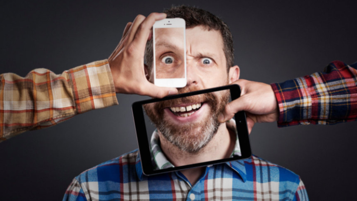 Dave Gorman gawps to camera, with a photo of his eye on a smartphone held up in front of his eye and photo of his mouth on a tablet held up in front of his mouth. Gorman and both hands holding the devices are in different checkered shirt