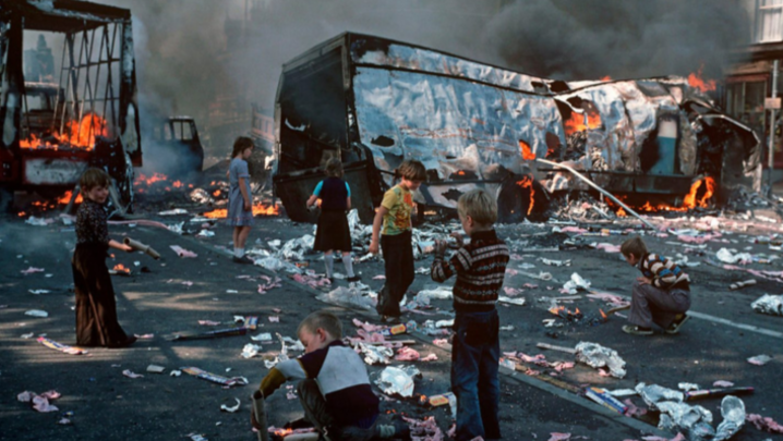 Seven children pick through the partially burning wreckage of a bus and other large vehicle. Detritus, also ablaze in places, litters the street