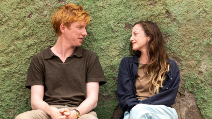 Alice and Jack sit next to each other, looking intently into each other's eyes, in front of a green wall