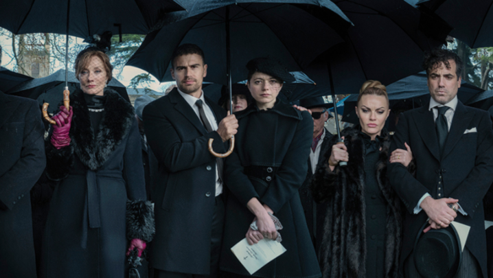 A group of people stand in black tie formalwear holding umbrellas, looking sombre