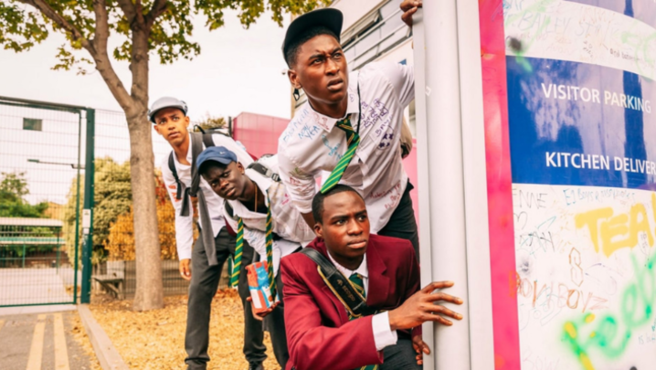 Characters from “Grime Kids” – Junior, Dane, Bishop and Bayo – hide behind a sign wearing school uniforms, which are covered in writing in coloured felt tip pen.