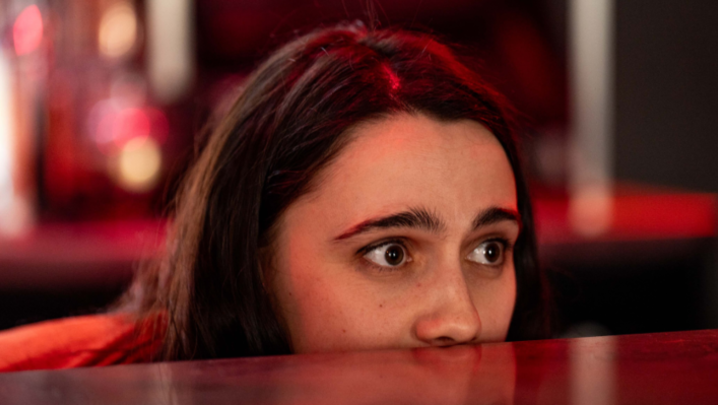 Kat Sadler, in character as Josie, peers out from over a bar, only the top half of her face visible