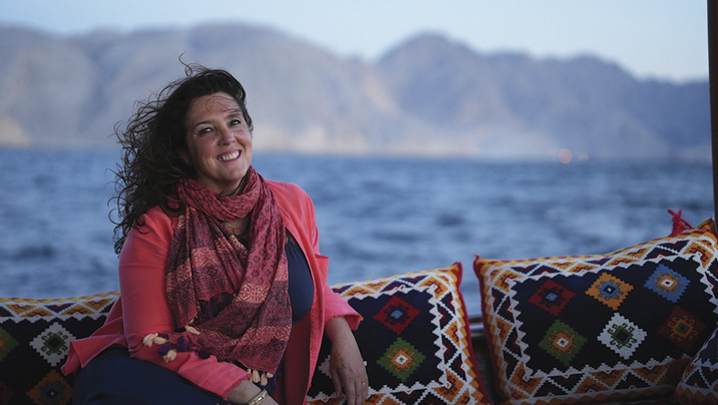 Bettany Hughes is pictured on an outdoor sofa overlooking a mountainous bay